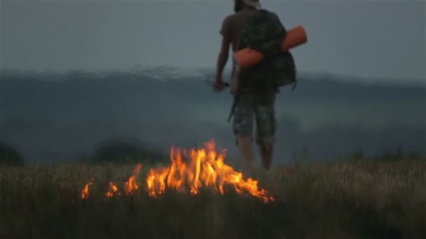 Vousatý Turistický batoh nedaleko hořícího pole. Muž v klobouku a cestovatel s batohem na pozadí plamenů. — Stock video