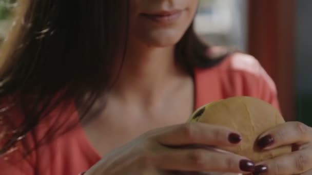 Retrato de una chica de cerca con una hamburguesa en la mano. Una joven mujer guapa comiendo una hamburguesa en un café . — Vídeos de Stock