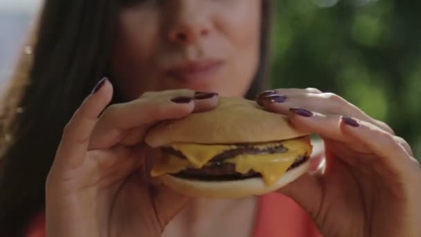 Retrato de una chica de cerca con una hamburguesa en la mano. Una joven mujer guapa comiendo una hamburguesa en un café . — Vídeo de stock