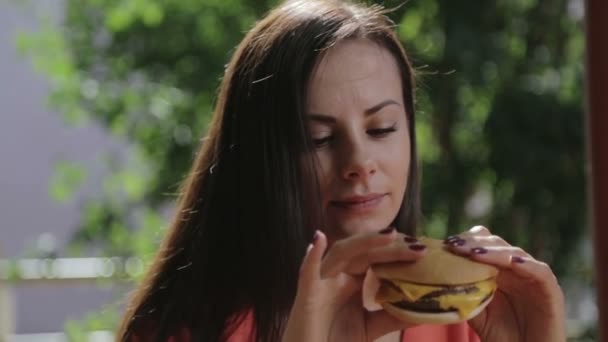 Ritratto di una ragazza primo piano con un hamburger in mano. Una giovane bella donna che mangia un hamburger in un caffè . — Video Stock