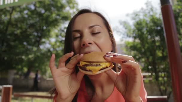 Retrato de una chica de cerca con una hamburguesa en la mano. Una joven y bonita mujer comiendo una hamburguesa en un café. Comida, Comida Rápida, Nutrición . — Vídeo de stock