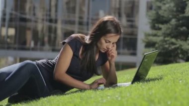 Bir dizüstü bilgisayar ile parkta genç güzel kadın. Güzel esmer çimenlerin üzerine oturmuş bir dizüstü bilgisayarda yazarak. İnsan, doğa, teknoloji. 