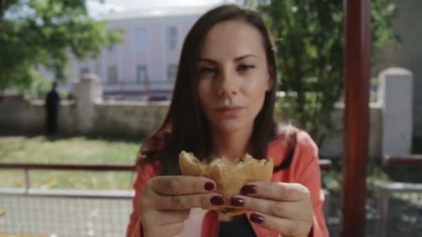 Portré egy lány zár-megjelöl egy hamburger a kezében. Egy fiatal csinos nő, eszik egy hamburgert, egy kávézóban. Élelmiszer, Fast Food, táplálkozás. — Stock videók