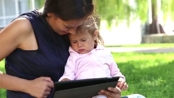 Madre enseña a una niña pequeña.Mamá enseña a un niño a jugar en la tableta.Madre joven con el bebé en el parque jugando en la computadora portátil . — Vídeos de Stock