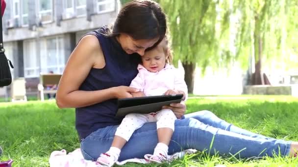 Madre enseña a una niña pequeña.Mamá enseña a un niño a jugar en la tableta.Madre joven con el bebé en el parque jugando en la computadora portátil . — Vídeos de Stock