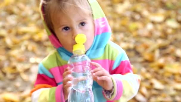 Barnet dricker mineralvatten. Lilla flickan är dricksvatten i naturen. Lilla flickan i ljusa kläder är dricksvatten från en flaska i höst park. Känslor barnet närbild. — Stockvideo