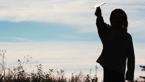 Silhouet van een jongen speelt met een papieren vliegtuigje. Eenzame jongen buiten spelen. Het kind begint vlak op hemelachtergrond. — Stockvideo