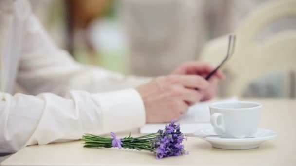 Mans mão com um close-up de uma xícara e um buquê de lavanda. Mão de homem com óculos no fundo do livro. Mão, livro, copo, flor , — Vídeo de Stock