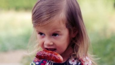 Beyaz küçük kız sosis mangal yiyor. Çocuk bir kız doğada yiyor. Emotsonalny çocuk sağlıklı gıda iştah açıcı yiyor. Bir yemek için bir çocuk portresi.