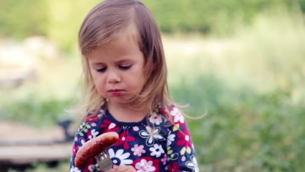 Une petite fille caucasienne mange un barbecue à saucisses. L'enfant mange une fille dans la nature. Emotsonalny enfant mange des aliments sains appétissants. Portrait d'un enfant pour un repas . — Video