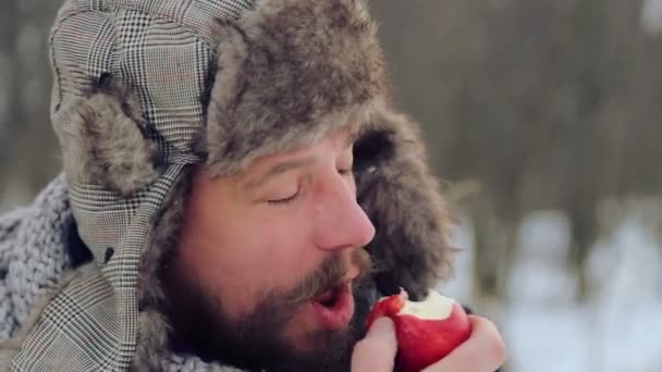 Porträtt av en skäggig man med ett äpple på vintern. En ung skäggig man äter ett äpple på vintern. Den skäggiga mannen äta ett äpple till kameran på vintern. — Stockvideo