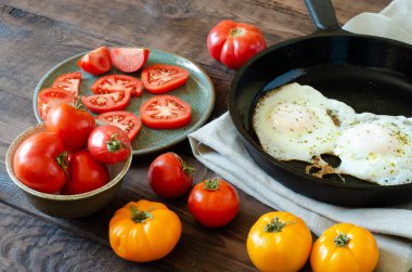 Tomatoes in bowl and sliced tomatoes on plate. Fried eggs on frying pan on linen and wooden background.
