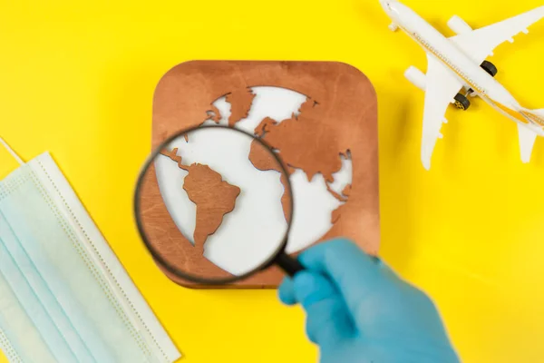 Plane model, face mask, loupe in hand and earth model on a yellow background. Hands in gloves. Flight impact of coronavirus (COVID-19) concept