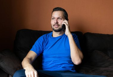 Sakallı yakışıklı adam, cep telefonuyla konuşuyor, evdeki kanepede oturuyor..