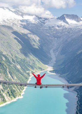 Tourist with red hoody sit on swing bridge looking at glacier reservoir in Austria zillertal Alps clipart