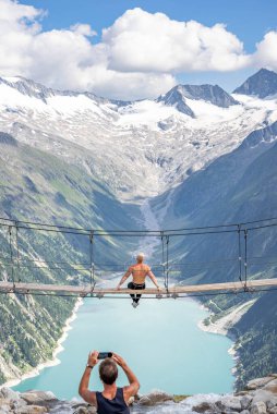 Olpererhutte, Austria - Aug 7, 2020: Man take photo of half naked man on swing bridge facing glacier clipart