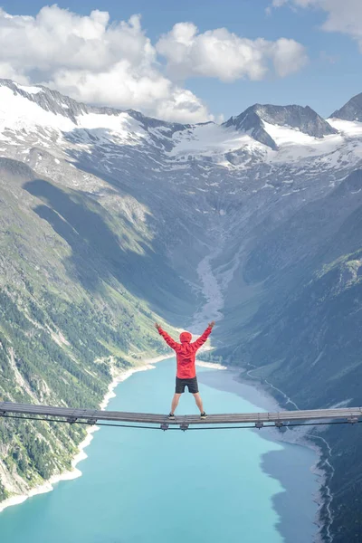 Turista s červenou kapucí sedí na houpacím mostě a dívá se na ledovcovou nádrž v rakouských zillertálních Alpách — Stock fotografie