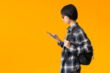 Asyalı genç kız akıllı telefon kullanıyor ve sırt çantası taşıyor, sarı arka planda izole edilmiş.