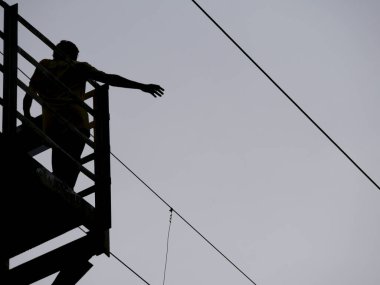 Bir adam inşa edilmiş köprü siluetinde dururken sağ elini tele doğru uzatıyor.
