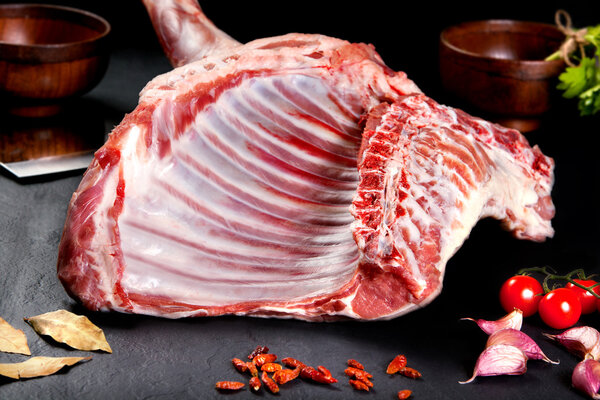 Fresh and raw meat. Whole piece of uncooked ribs and chops to grill, roast barbecue. Black stone background with tomato and pepper