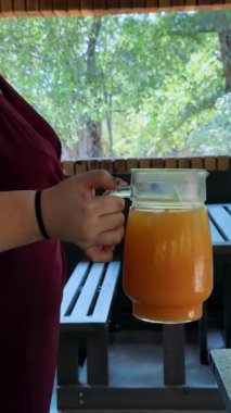 Portakal suyu gri arka plandaki bir bardağa dökülür. Bir el masaya bir sürahi portakal suyu koyar.