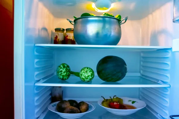 Shelves of an open frige refrigerator with different food, cooked food in pot and plastic container.