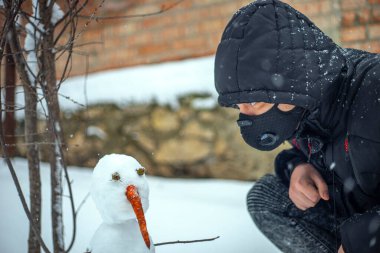 Koruyucu maskeli bir adam yoğun kar yağışı sırasında kardan adamla oturuyor.