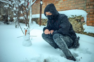 Koruyucu maskeli bir adam yoğun kar yağışı sırasında kardan adamla oturuyor.
