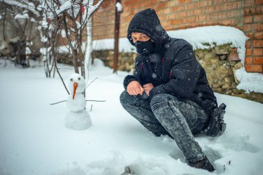 Koruyucu maskeli bir adam yoğun kar yağışı sırasında kardan adamla oturuyor.