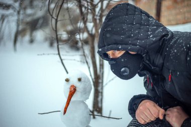 Koruyucu maskeli bir adam yoğun kar yağışı sırasında kardan adamla oturuyor.
