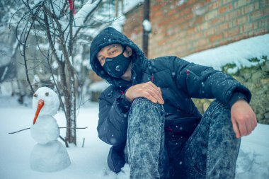 Koruyucu maskeli bir adam yoğun kar yağışı sırasında kardan adamla oturuyor.