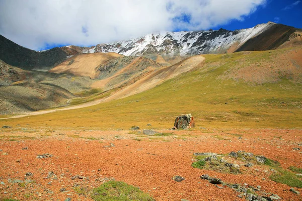 Güneşli vadi manzarası, karlı dağ tepeleri ve bulutlu gökyüzü.