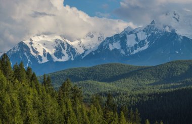 Mountain landscape, snow-capped peaks and trees. Summer evening, cloudy sky.