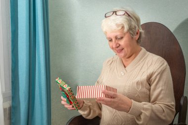 Gözlüklü yaşlı bir kadın evinde bir sandalyede oturuyor, bir kutuyu hediyeyle açıyor ve gülümsüyor.