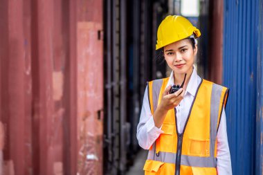 Young confident woman engineer smiling and using radio communication and wearing yellow safety helmet and check for control loading containers box from Cargo freight ship for import and export