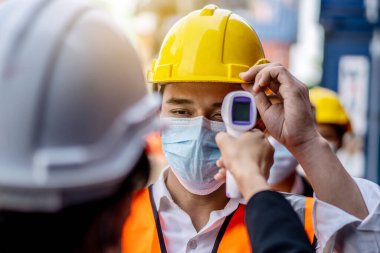Female foreman wearing a mask measures the temperature for a worker in a safety suit. They stood and waited for the measurement with a non-contact infrared thermometer to prevent covid virus