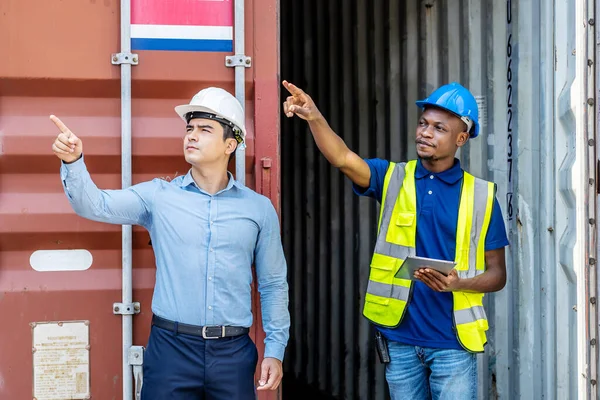 Port manager and a colleague tracking inventory while standing point to position loading Containers box from Cargo freight ship at Cargo container shipping on a large commercial shipping dock