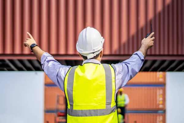 Rear view of engineer in reflective clothing with safety helmet is working and checking at containers cargo on shipping port area at harbor