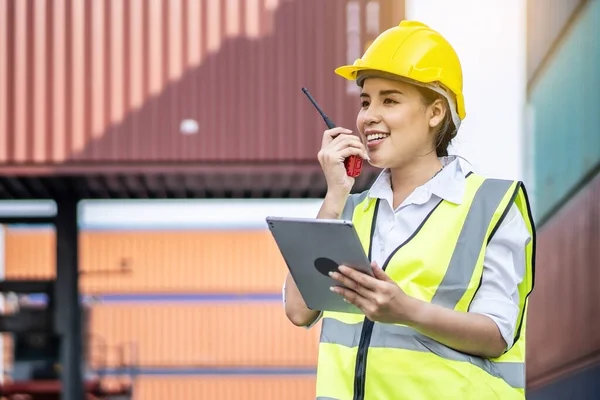 Young asian female engineer or manager holding a Tablet and use of radio communication to control container load in industrial transport and logistics concepts