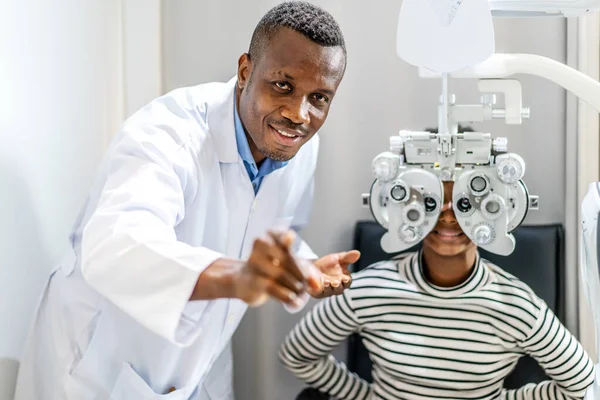 Optician doing optometry eye exam for black african american teen girl patient. Male optometrist with phoropter while examining patient.