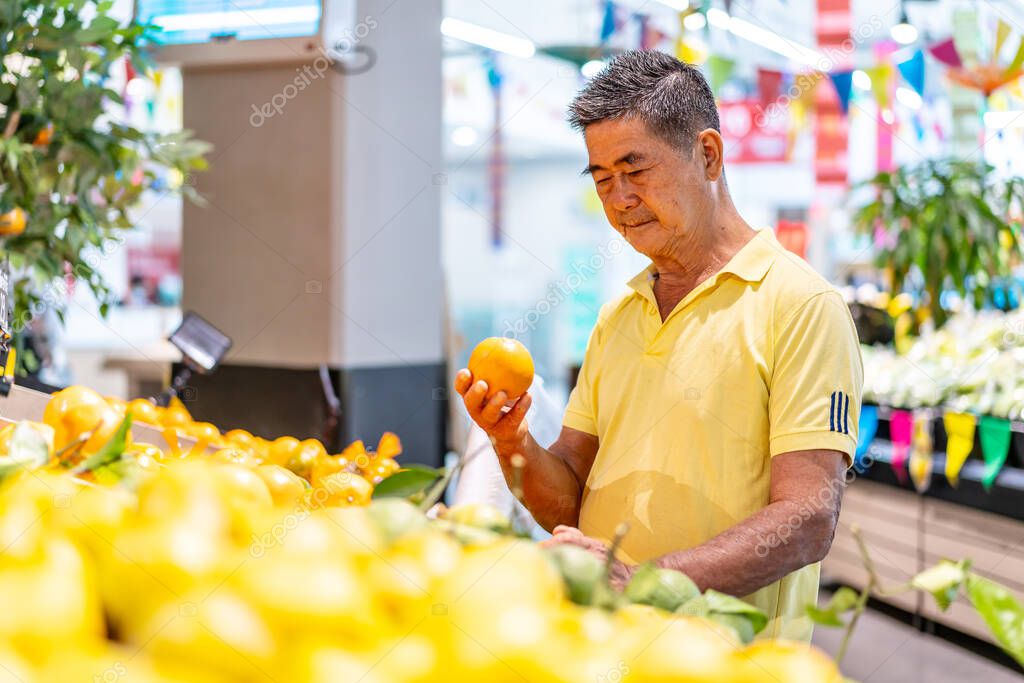 Old senior asian man looking at tangerine while enjoying grocery shopping at in the supermarket