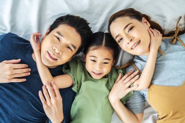Mutlu Asyalı genç anne, baba ve kızının yatak odasında baş başa yatarken kameraya bakıp gülümsemelerinin en üst görüntüsü.