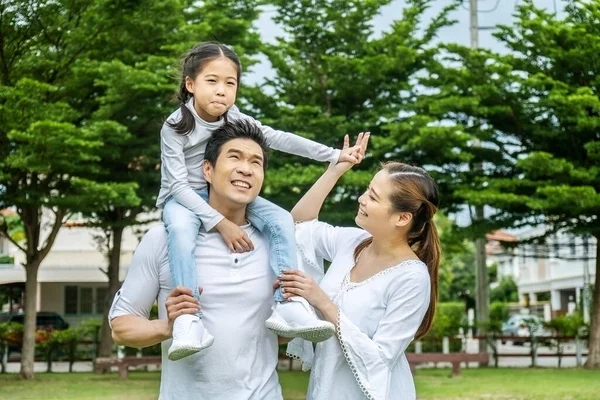 Portrait of asian happy family of three having fun together in park,  little daughter sitting on father back and mother smile walking and having fun moments good time in summer park at home