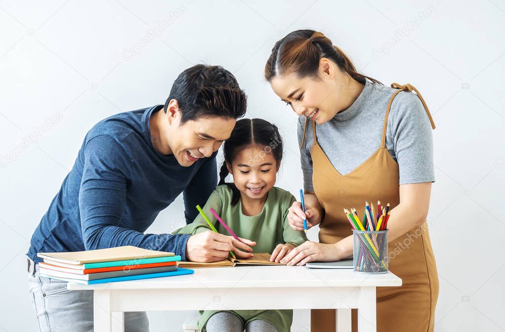 Asian young mother and father with little daughter sit at desk learning and writing in book with pencil making homework at home.Education concept.