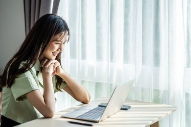 Evde dizüstü bilgisayar kullanan gülümseyen Asyalı genç bir kadın, güzel bir kız alışveriş yapıyor veya sosyal ağda sohbet ediyor, eğleniyor, film izliyor, bilgisayar projesinde serbest çalışıyor.