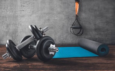 Ahşap zeminde halterler, yoga minderi ve arkasında TRX kayışları var..