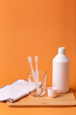 Bardakta beyaz diş fırçaları, gargara ve turuncu arka planda tahta masanın üzerinde yüz havlusu. Ağız bakımı, sakız ve diş sağlığı. Sabah rutini ve diş bakımı.