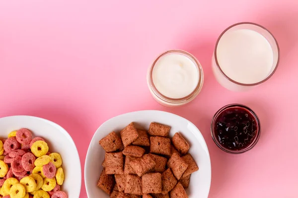 Kuru kahvaltı ve süt. Kuru tatlı kahvaltı - mısır halkaları, çikolatalı mısır yastıkları, süt, yoğurt ve pembe arka planda bal. Hızlı ve besleyici bir kahvaltı.