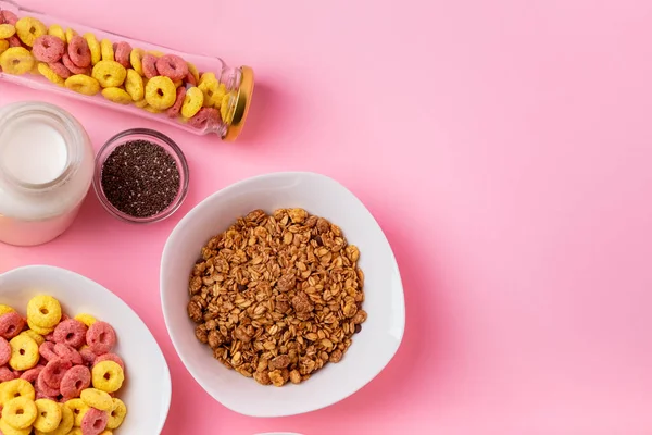 Kuru kahvaltı ve süt. Kuru tatlı kahvaltı - mısır halkaları, çikolatalı mısır yastıkları ve granola, süt, yoğurt ve bal pembe arka planda. Hızlı ve besleyici bir kahvaltı.