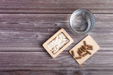 Tahta kutuda beyaz renkli haplar, vitaminler ve mineraller, beslenme takviyeleri ve ahşap arka plandaki ahşap tepside yukarıdan gelen bir bardak su. Vitaminler ve kırsal tarzda beslenme takviyeleriyle güzel bir kompozisyon. Vitaminler ve mineraller.
